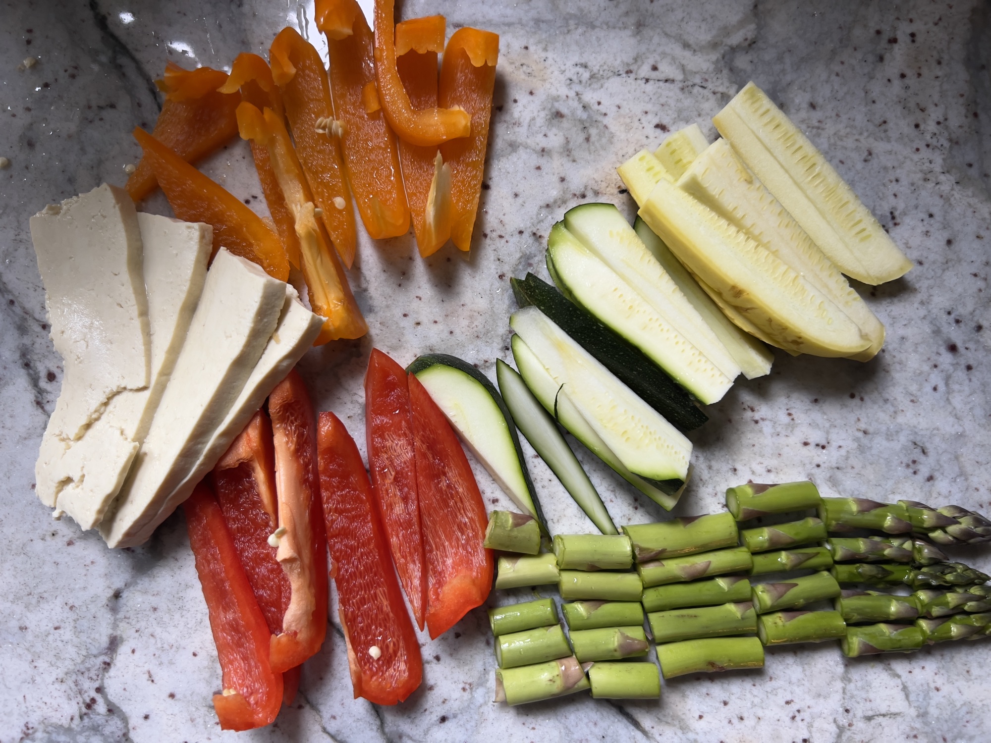 Tofu, bell peppers, zucchini, and asparagus prepped on a marble counter